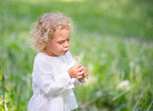 A blonde girl in a light dress blows on a white dandelion flower in the hope that fluffs will fly from it