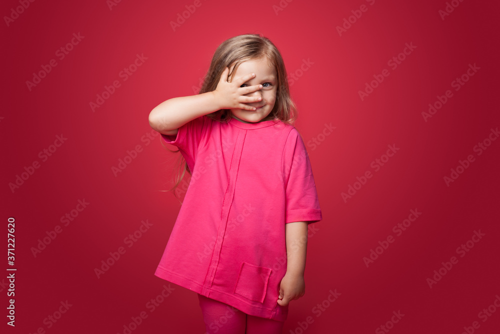Smiling blonde caucasian girl is covering her eyes with a palm posing on a red studio wall Smiling blonde caucasian girl is covering her eyes with a palm posing on a red studio wall
