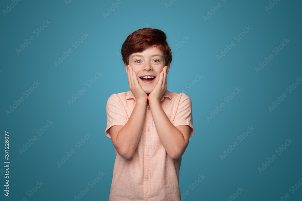 Surprised caucasian boy with red hair covering cheeks with palms is smiling on a blue studio wall Surprised caucasian boy with red hair covering cheeks with palms is smiling on a blue studio wall