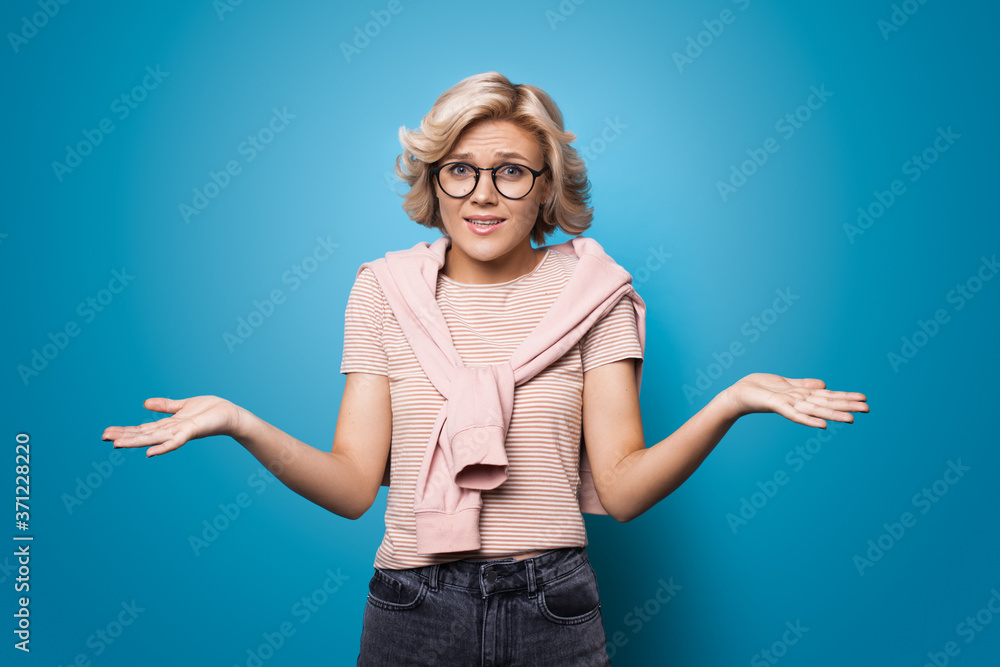 Confused blonde woman gesturing with palms at looking at camera through glasses on a blue studio wall