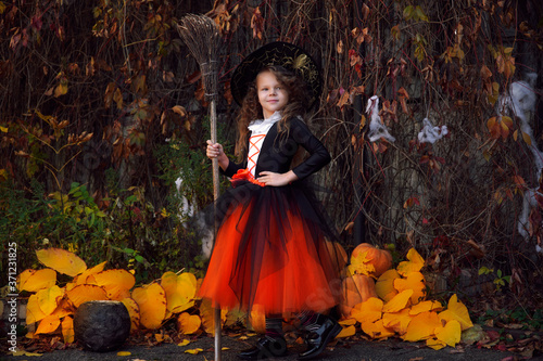 a girl in an orange skirt and a black hat on Halloween near pumpkins with a broom on a background of yellow leaves                 