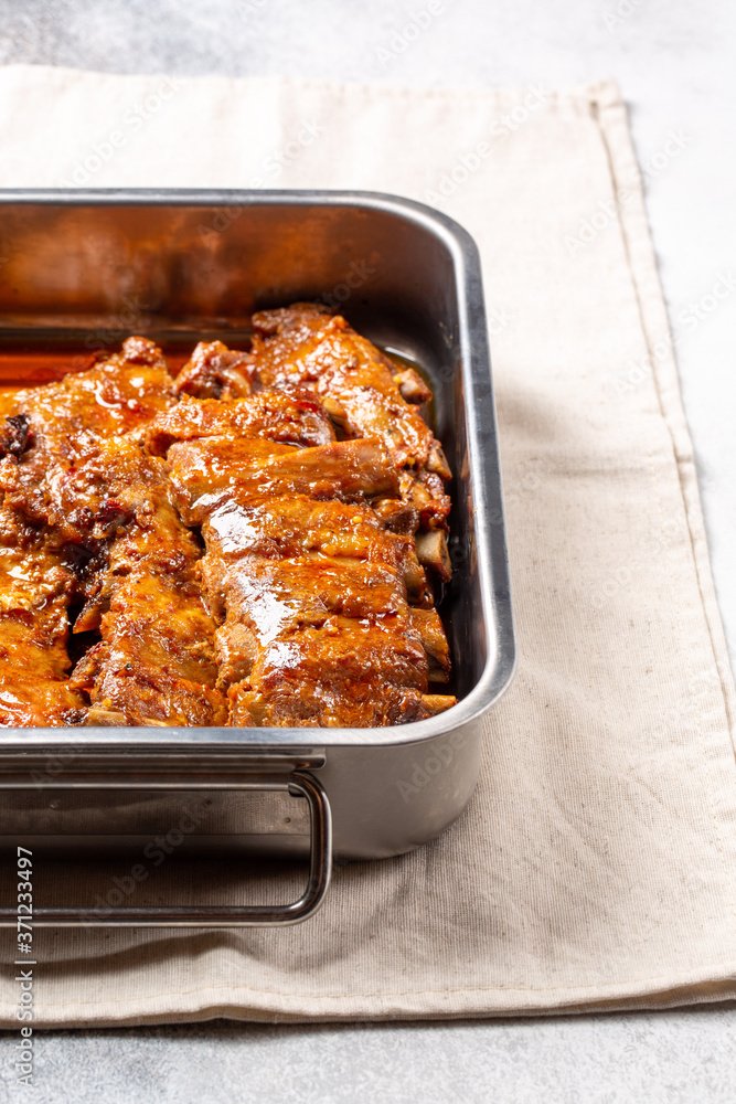 Oven baked pork ribs in spicy sauce in metal baking dish on concrete background.
