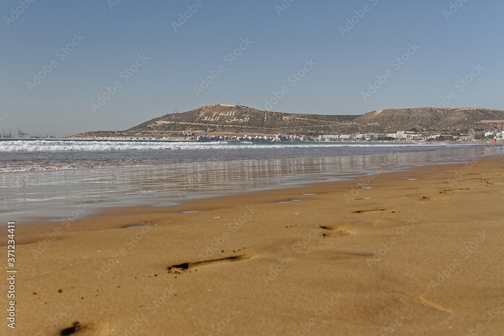 Agadir, Morocco.Feb.3,2019:Beach with tourist walking and Moroccan motto on the mountain. Writing on the hillside meaning, God, Country, King. People walking on the beach footsteps in the sand.