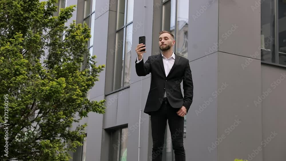 Caucasian handsome young businessman in suit standing at street at business center and talking via videochat on smartphone. Man videochatting on webcam of mobile phone. Video call. Caucasian handsome young businessman in suit standing at street at business center and talking via videochat on smartphone. Man videochatting on webcam of mobile phone. Video call.