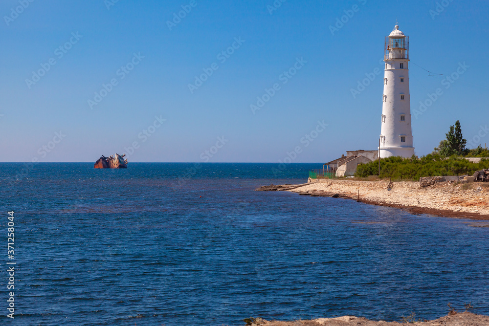 A white stone lighthouse stands on the seashore. Not far from it in the sea is an old half-sunken ship