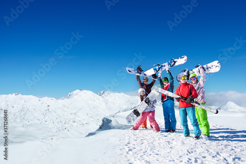 Large group of young snowboarders lift snowboards over head standing on top of the alpine mountain