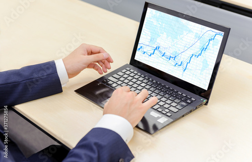 ノートパソコンでモニター上の株価チャートを見るビジネスマン
