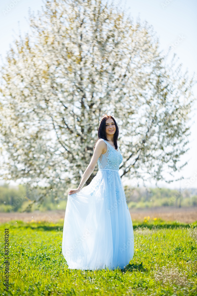 Portrait of a girl in a light summer blue long dress adorned in her hair against a blossoming tree.