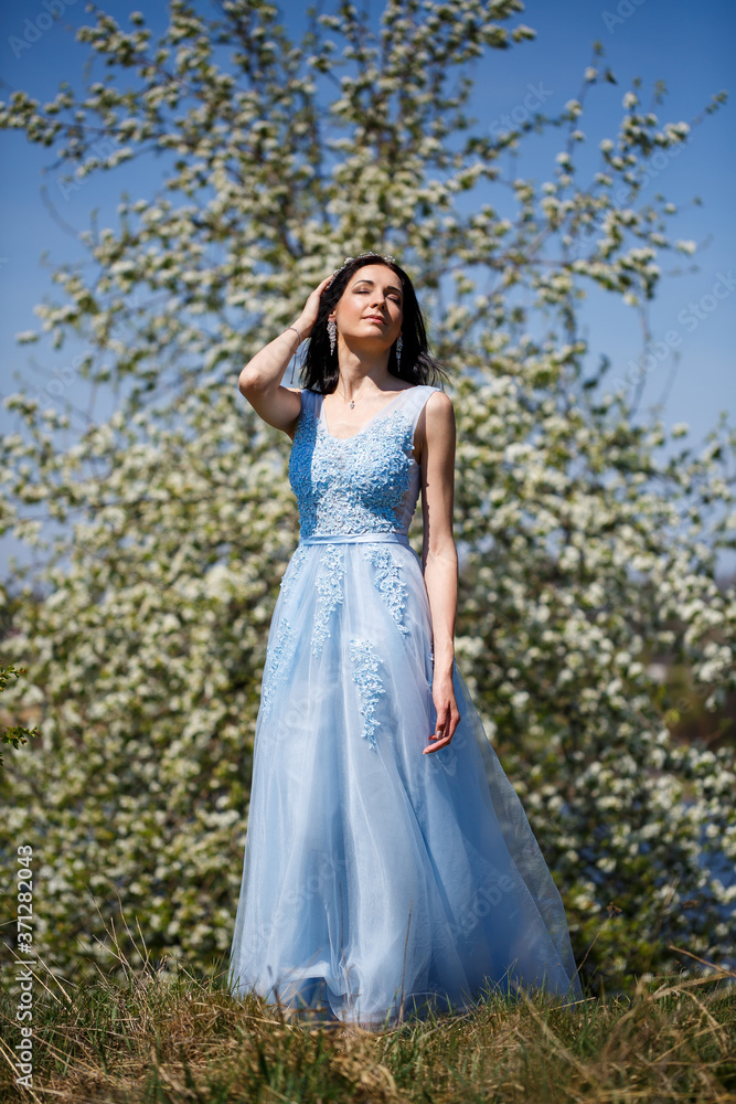 Portrait of a tender girl in a blue long dress with tulle under blooming cherry with a smile on her face on a sunny warm summer day