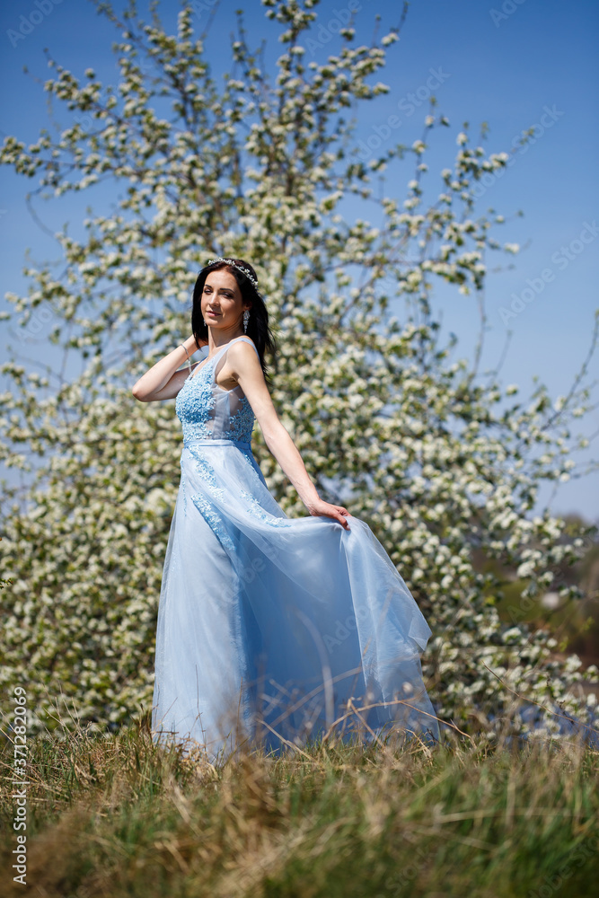 Portrait of a tender girl in a blue long dress with tulle under blooming cherry with a smile on her face on a sunny warm summer day