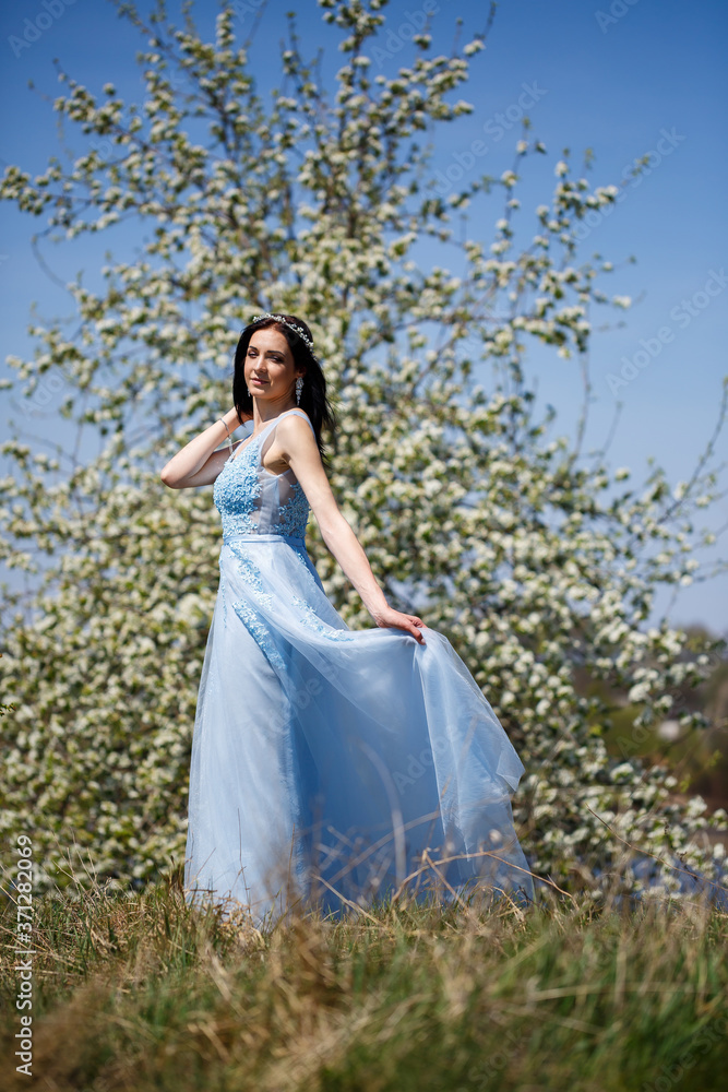 Portrait of a tender girl in a blue long dress with tulle under blooming cherry with a smile on her face on a sunny warm summer day Portrait of a tender girl in a blue long dress with tulle under blooming cherry with a smile on her face on a sunny warm summer day