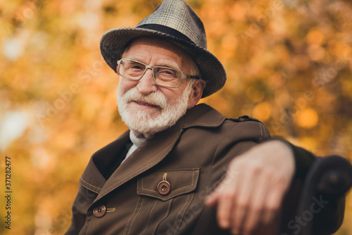 Closeup photo of funny glad old pensioner grey haired grandpa central park walk enjoy sunny day warm weather sit bench break pause wear stylish autumn jacket hat specs colorful street outside