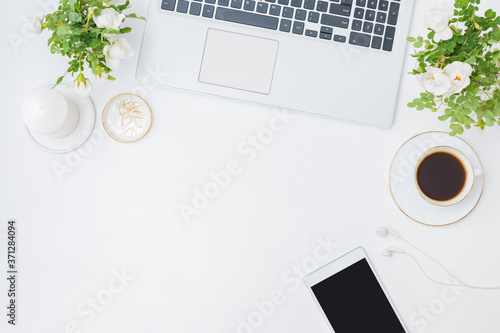 Flat lay blogger or freelancer workspace with a laptop, cup of coffee, small flowers and green leaves, office supplies on a light background