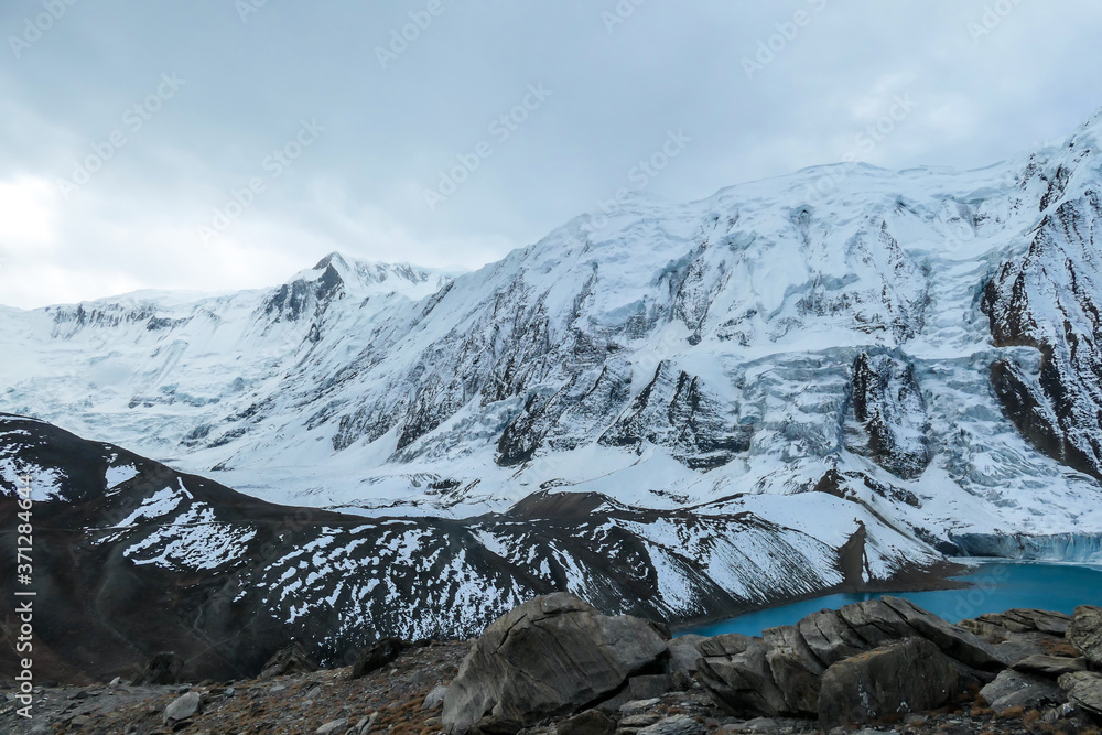 Stormy weather in snow capped Himalayan peaks along Annapurna Circuit in Nepal. Barren and sharp slopes. Mountains are partially shrouded with clouds. There is a turquoise Tilicho Lake on the side.