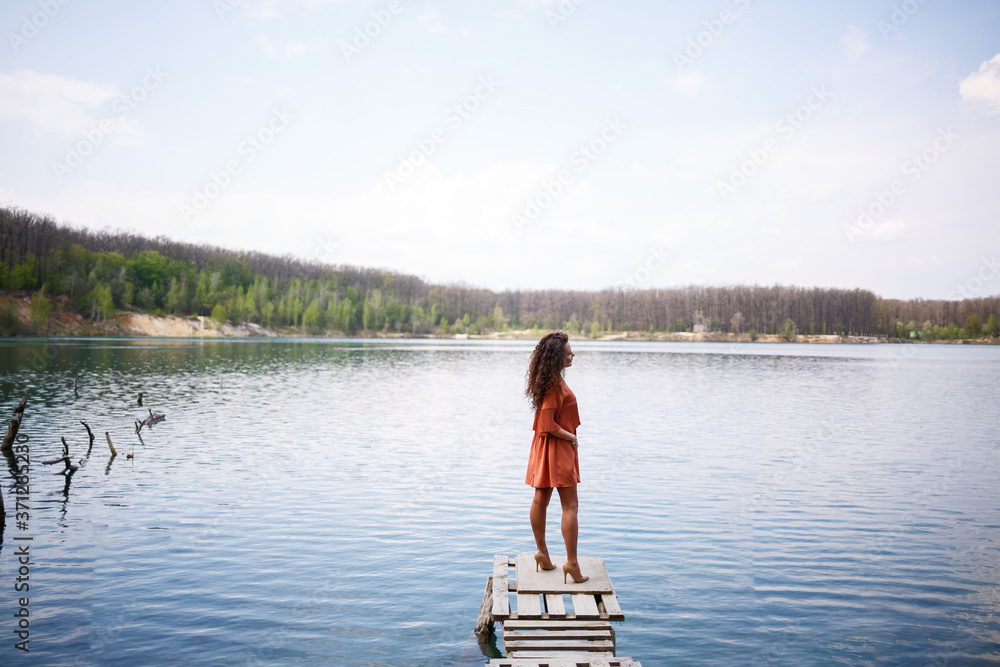 Wooden masonry. A girl with long wavy curly hair in an orange guipure dress and shoes on nature, in a forest against a lake, stood near trees and bushes. Young woman smiles and enjoys life Wooden masonry. A girl with long wavy curly hair in an orange guipure dress and shoes on nature, in a forest against a lake, stood near trees and bushes. Young woman smiles and enjoys life