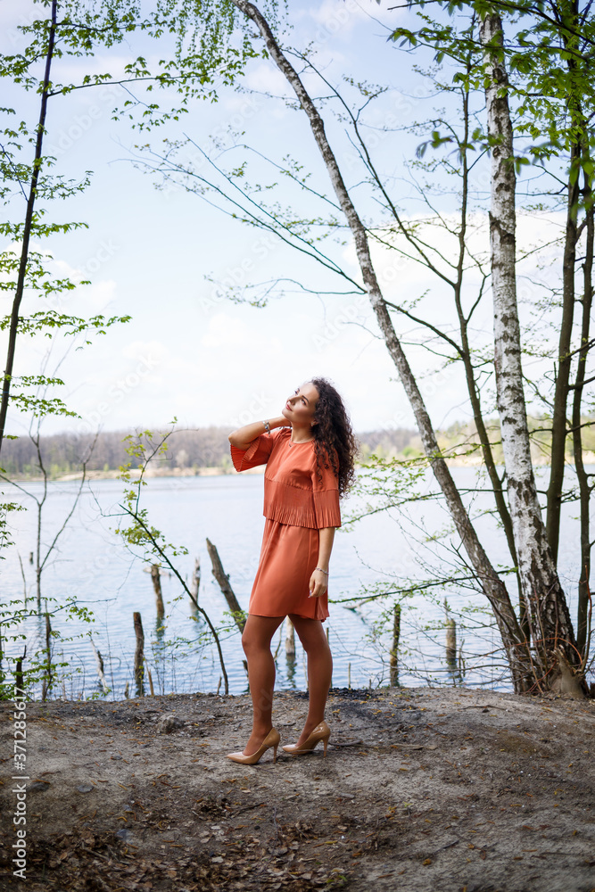 Portrait of a young woman with a smile on her face, happy emotions. A girl with long wavy curly hair in a dress in the summer on nature, in a forest on a lake, stood near a road and bushes Portrait of a young woman with a smile on her face, happy emotions. A girl with long wavy curly hair in a dress in the summer on nature, in a forest on a lake, stood near a road and bushes