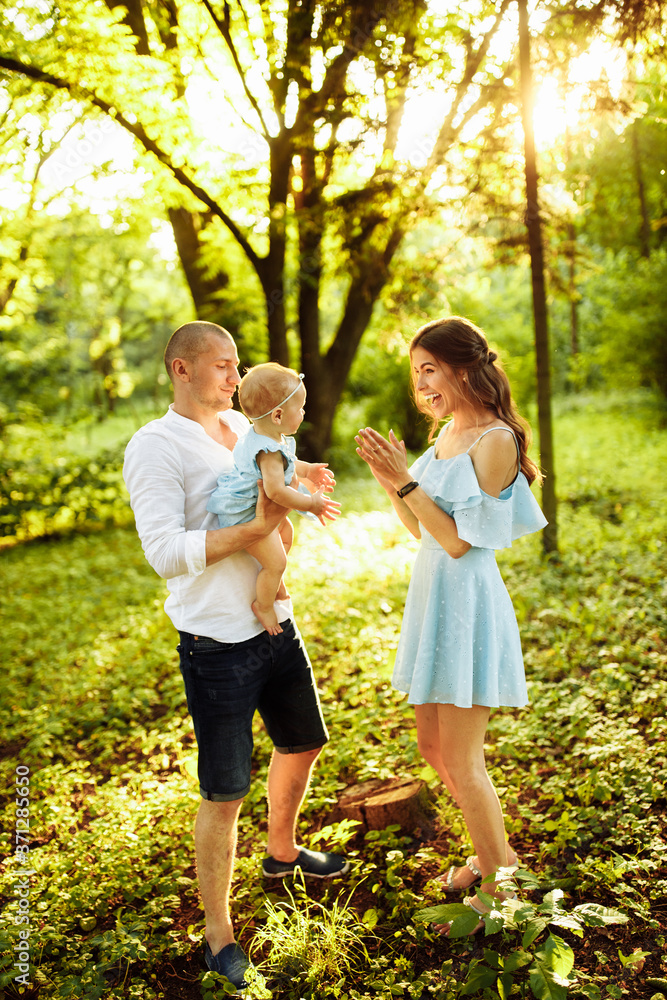 Fototapeta premium Young family walking at the park, caring parents play with little daughter, smiling, loving dad hold adorable baby girl in arms, enjoy weekend outdoors, parenthood concept