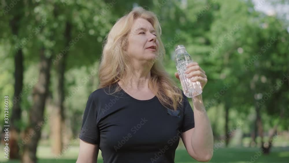 Camera approaches to satisfied mid-adult woman drinking refreshing water from bottle in summer park. Portrait of confident happy Caucasian sportswoman having break in training outdoors. Lifestyle. Camera approaches to satisfied mid-adult woman drinking refreshing water from bottle in summer park. Portrait of confident happy Caucasian sportswoman having break in training outdoors. Lifestyle.
