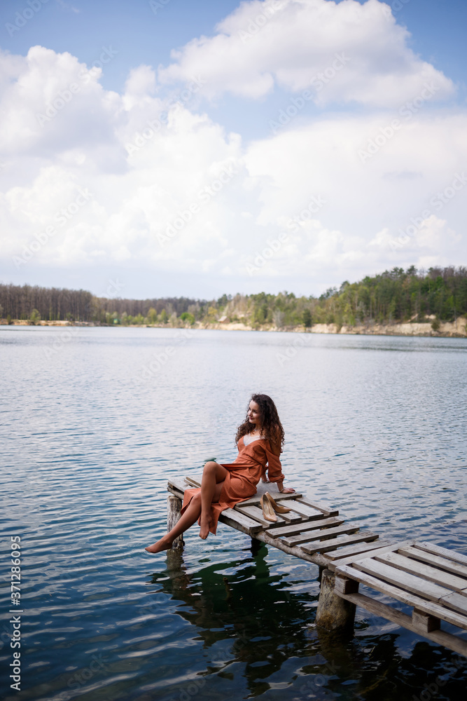 A girl with long wavy curly hair in a long guipure dress barefoot in the summer in a forest on a lake at sunset standing on a pantone on a wooden pier bridge. Summer sunny day A girl with long wavy curly hair in a long guipure dress barefoot in the summer in a forest on a lake at sunset standing on a pantone on a wooden pier bridge. Summer sunny day