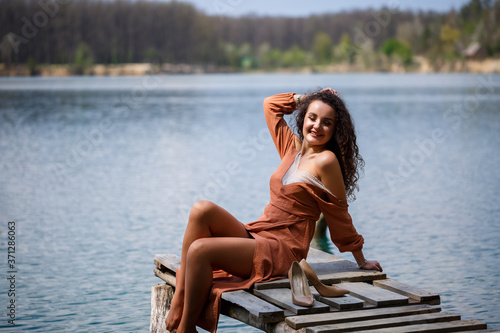 A girl with long wavy curly hair in a long guipure dress barefoot in the summer in a forest on a lake at sunset standing on a pantone on a wooden pier bridge. Summer sunny day photo