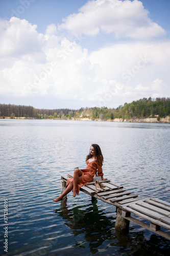 A girl with long wavy curly hair in a long guipure dress barefoot in the summer in a forest on a lake at sunset standing on a pantone on a wooden pier bridge. Summer sunny day