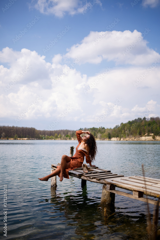 Young woman with long wavy curly hair in a long guipure dress barefoot in the summer in a forest on a lake at sunset standing on a pantone on a wooden pier bridge. Summer sunny day Young woman with long wavy curly hair in a long guipure dress barefoot in the summer in a forest on a lake at sunset standing on a pantone on a wooden pier bridge. Summer sunny day