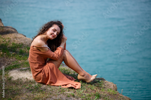 A young woman with curly hair and a smile on her face sits in a green meadow on a background of blue lake. Warm summer day, happy girl, emotions of joy