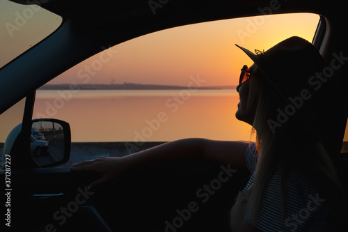 A young woman looks out the car window at the sunset on the sea.