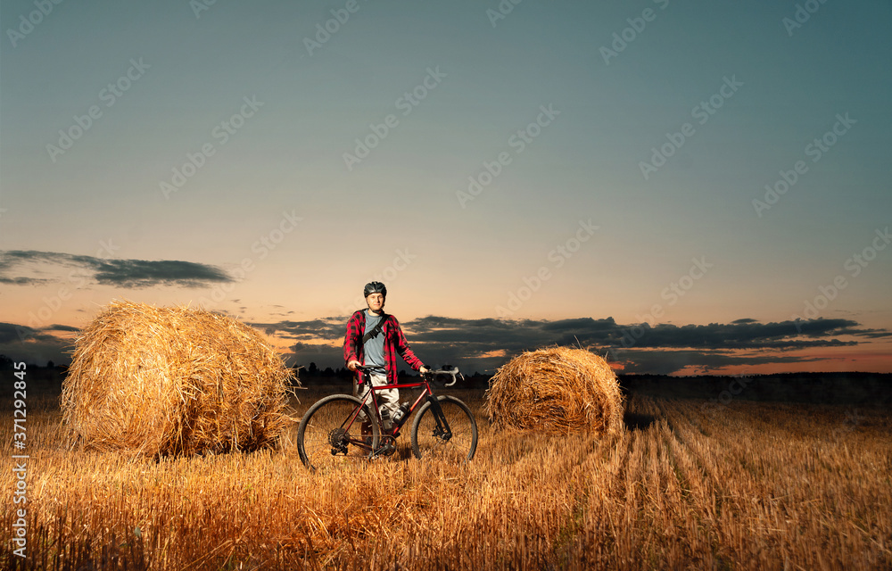 Obraz premium A cyclist in a helmet with bicycle stands in a field with bales of straw at sunset. Active and sporty lifestyle concept.