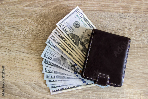 Money dollars lie under a leather wallet on a wooden table. Wallet lies on money dollars in the office on the table.