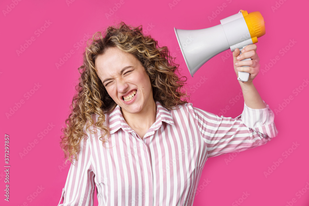 Naklejka premium A young woman holds a screaming megaphone in her hands, which is aimed at her face. Isolated pink background.