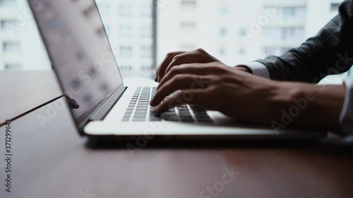 Close-up of hands of unrecognizable business man typing on laptop keyboard while working at office desk on background of large panoramic window. Camera moves smoothly towards the keyboard. photo