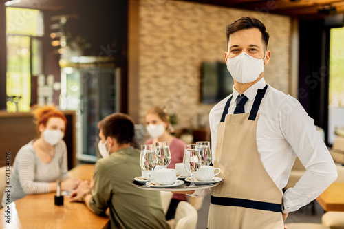Happy waiter serving coffee to his customers while wearing protective face mask in a cafe