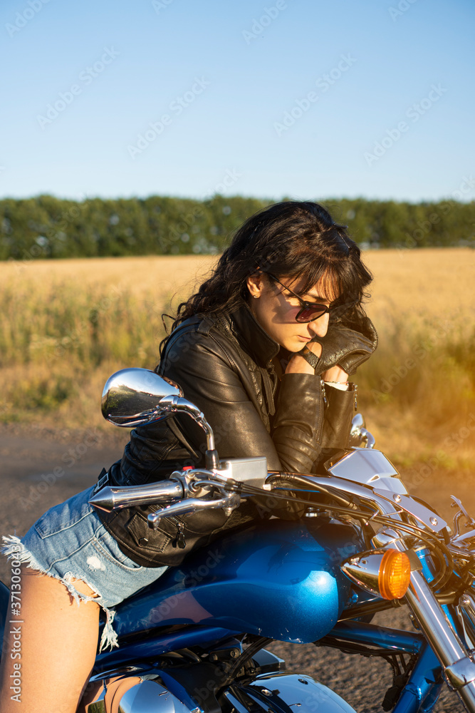 Fashionable biker girl sits on the motorcycle. She stopped on the rural country road behind the field.
