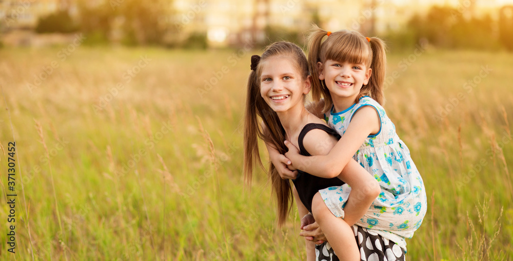 Fototapeta premium Happy child girl holding little sister in arms on back. Happy childhood together in summer time