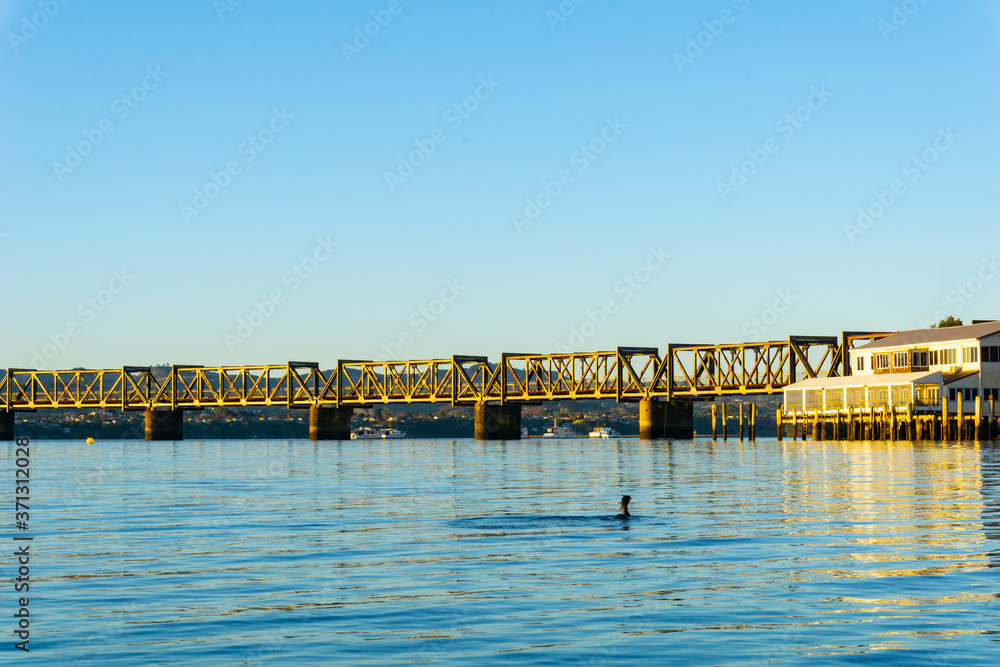 Tauranga Railway bridge catches glow from sunrise in distance across  blue calm water