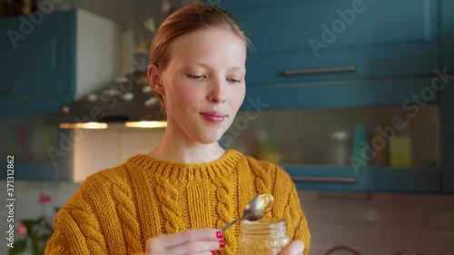 Close up young woman in kitchen eating honey from jar with spoon and enjoying meal. Attractive woman eating healthy food spoon