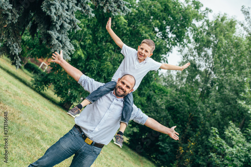 a bearded man rolls on the shoulders of a teenage boy in a park. Dad and son play airplanes in nature,