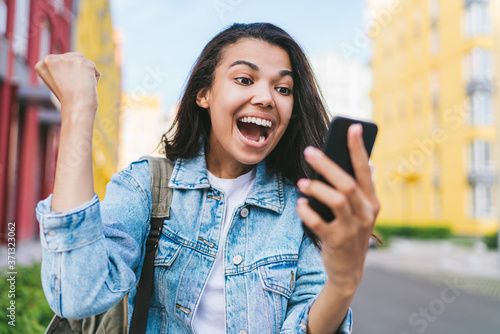 Portrait of cute dark skinned girl showing sincere happiness about victory at bookmaker's website celebrating and clenching her fist in winner's gesture and screaming yes