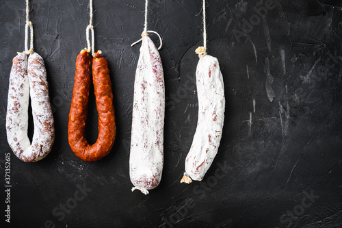 Spanish salami, fuet and chorizo sausages hang from a rack on black textured background with copy space
