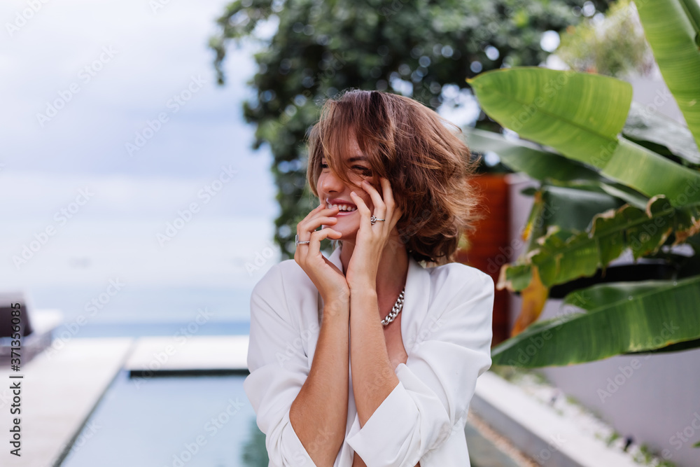 Fashion portrait of woman at tropical luxury villa wearing white stylish blazer, rings, earings on background of tropical leaves and swimming pool. Fashion portrait of woman at tropical luxury villa wearing white stylish blazer, rings, earings on background of tropical leaves and swimming pool.