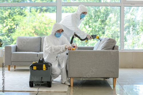Workers in biohazard costume removing dirt from sofa in house