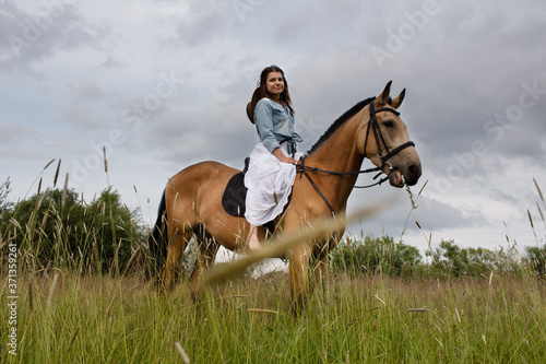 Young cow girl walking horse in green field. People and animal portrait. Horseback riding view. Active weekend