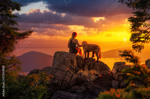 Adventurous Girl is hiking with a dog on top of St. Mark's Mountain during a dramatic summer sunset. Located in West Vancouver, British Columbia, Canada.