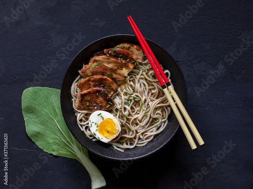 A bowl of ramen chicken soup with egg and sticks on black background.