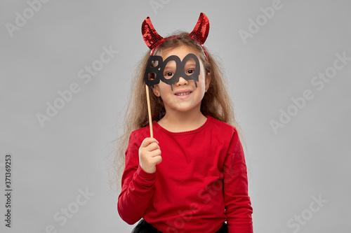 halloween, holiday and childhood concept - smiling girl in costume with red devil's horns holding party accessory over grey background