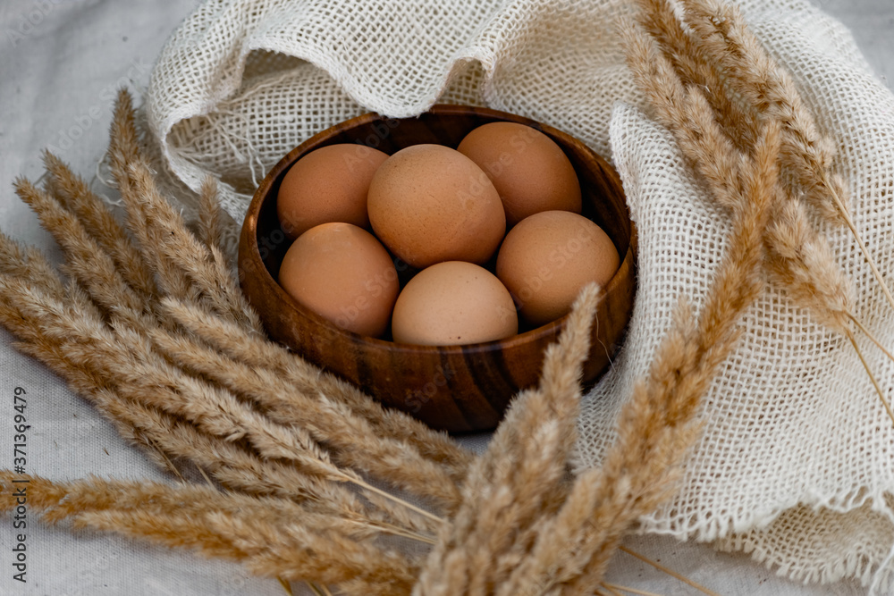Chicken eggs in a deep wooden plate are laid out in a circle, next to dry twigs of Golden ears and dairy rustic natural fabric Chicken eggs in a deep wooden plate are laid out in a circle, next to dry twigs of Golden ears and dairy rustic natural fabric
