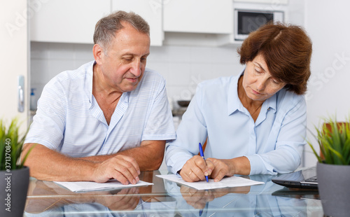 Image of smiling mature couple at table in home kitchen filling up documents