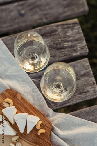 Two glasses of white wine and wooden plate with cheese and nuts during sunset time outside.