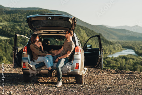 Young Traveler Couple on a Road Trip, Man and Woman Sitting on the Opened Trunk of Their Car Over Sunset © Romvy